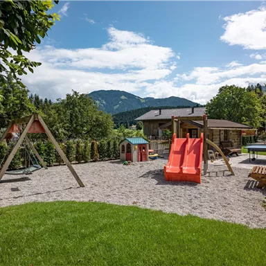 Ein schöner Spielplatz mit einer Rutsche und Schaukeln. Im Hintergrund sind Berge und ein Haus sichtbar.