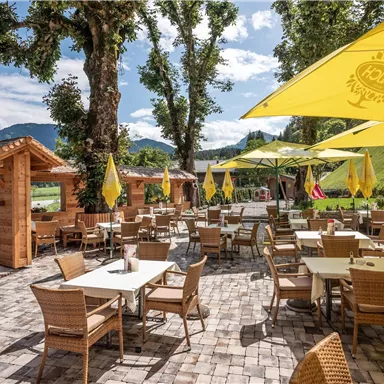 Ein gemütliches Restaurant im Freien mit Tischen und Stühlen. Gelbe Sonnenschirme bieten Schatten an einem sonnigen Tag.