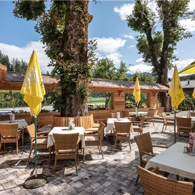 Ein gemütliches Restaurant im Freien mit Holzterrasse und Sesseln. Gelbe Sonnenschirme spenden Schatten unter blauem Himmel.