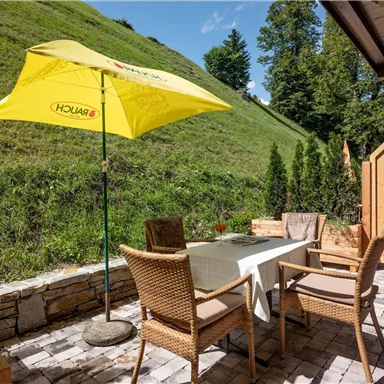 Ein einladender Außenbereich mit einem Tisch und Stühlen unter einem Gelben Schirm. Im Hintergrund sind grüne Hügel und Bäume zu sehen.