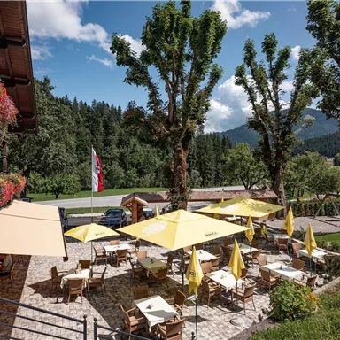 Ein einladendes Restaurant mit zahlreichen Tischen im Freien und gelben Sonnenschirmen. Im Hintergrund sind grüne Bäume und ein klarer Himmel zu sehen.