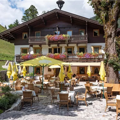 Ein gemütliches Gasthaus in den Bergen mit blühenden Fensterbänken. Die Terrasse ist mit gelben Schirmen und Holzmöbeln ausgestattet, ideal für Außenangebote.