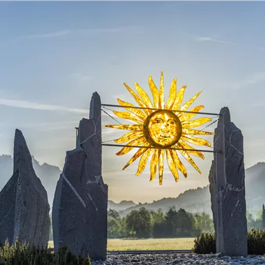 A sunny landscape featuring a large golden sun symbol placed between two stone sculptures. In the background, gentle mountains and a blue sky can be seen.