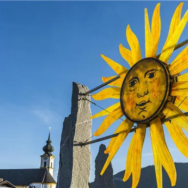 Ein strahlender Sonnensymbol mit einem Gesicht, umgeben von gelben Strahlen. Im Hintergrund sind ein Kirchturm und Felsen sichtbar.