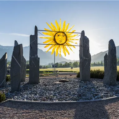 Ein beeindruckendes Steindenkmal mit einem großen, kunstvollen Sonnensymbol im Hintergrund. Die Szene ist von einer malerischen Berglandschaft umgeben.