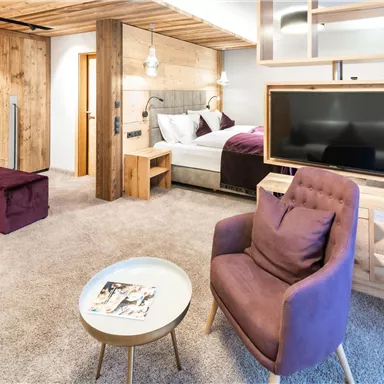 A modern hotel room with wooden details and a cozy atmosphere. There is a comfortable seating unit, a table, and a television.