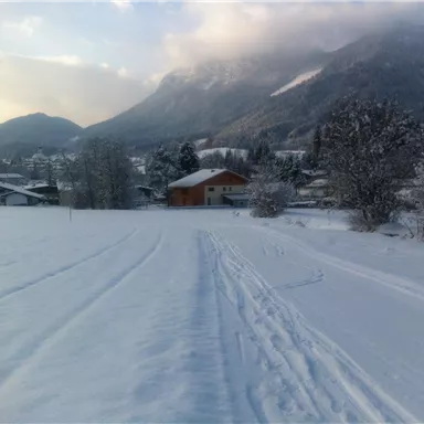 Eine verschneite Landschaft mit Hügeln im Hintergrund. Ein gemütliches Haus steht in der Nähe, umgeben von Bäumen.