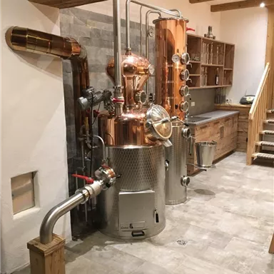 A copper still stands in a tastefully decorated room. The floor is tiled, and there are wooden shelves in the background.