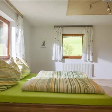 A cozy bedroom with a large bed and colorful pillows. Large windows provide plenty of daylight and a view of the greenery.