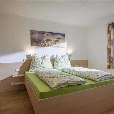 A cozy bedroom with two single beds and green bedding. The room features natural wood details and a window with curtains.