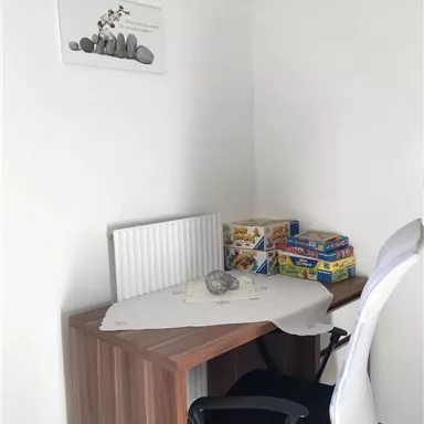 A simple workspace with a wooden table and a white chair. On the table is a tablecloth, a stone, and some games.