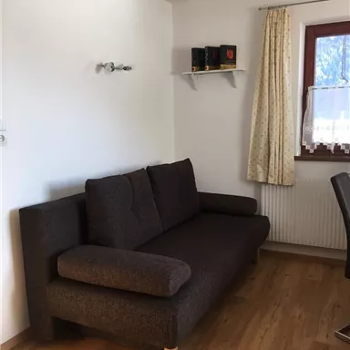 A cozy living room with a brown sofa and a window. On the wall, there is a shelf with decorations.