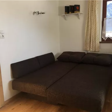 A simple room with a converted sofa and a window that lets in daylight. The walls are light, and there is a small shelf area.