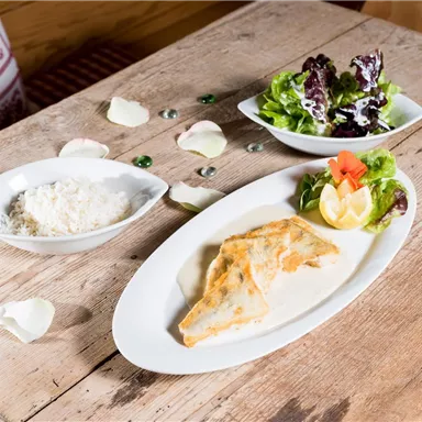 Ein köstliches Gericht mit einem Fischfilet in Sauce, serviert mit frischem Salat und einer Zitronenscheibe. Nebenbei liegt ein Schälchen mit Reis.