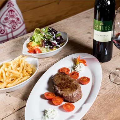 Ein köstliches Gericht mit Steak, frischen Tomaten, Pommes frites und einem bunten Salat. Dazu ein Glas Rotwein auf einem Holztisch.