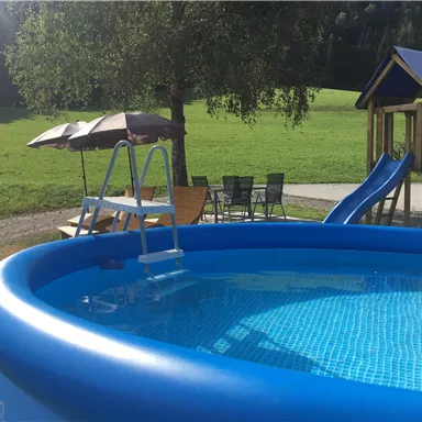 An inflatable pool with a ladder is in a green garden. Next to the pool, there are a slide, sun loungers, and umbrellas.