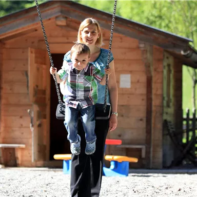 Eine Frau schwingt ein Kind auf einer Schaukel in einem Spielplatz. Im Hintergrund ist eine Holzhütte zu sehen.