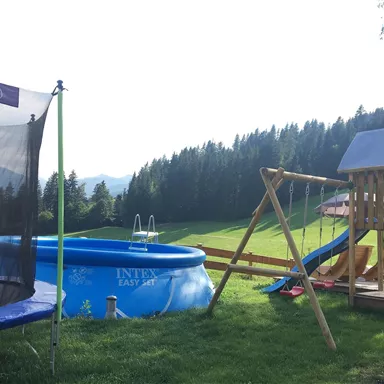 A play area with a trampoline, a swimming pool, and a playground. In the background, green meadows and trees can be seen.