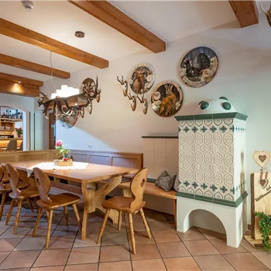 Ein gemütlicher Essbereich mit einem großen Tisch und Holzbänken. An den Wänden hängen dekorative Trophäen und ein traditioneller Ofen steht in der Ecke.