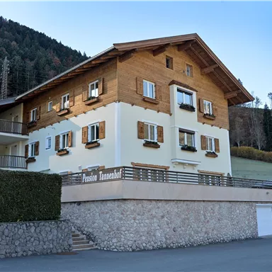 Ein charmantes, zweigeschossiges Haus mit Holzverkleidung und Fensterläden. Es liegt in einer malerischen Landschaft umgeben von Wäldern.