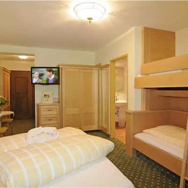 Ein gemütliches Hotelzimmer mit Doppelbett und Etagenbett. Im Hintergrund sind ein Fernseher und ein Schreibtisch zu sehen.