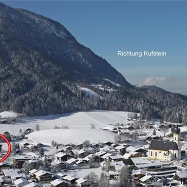 Eine verschneite Landschaft mit einem kleinen Dorf im Vordergrund. Im Hintergrund sind Berge und ein blauer Himmel zu sehen.
