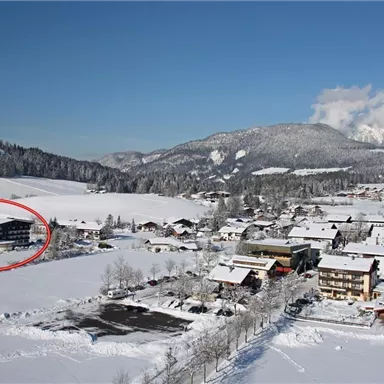 Eine verschneite Landschaft mit einem kleinen Dorf und Bergen im Hintergrund. Ein Gebäude ist im Vordergrund eingezeichnet.
