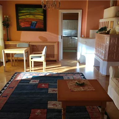 Ein gemütliches Wohnzimmer mit einem Teppich, einem Esstisch und Stühlen. Die Wände sind in warmen Farben gestrichen, und es gibt eine offene Küche im Hintergrund.