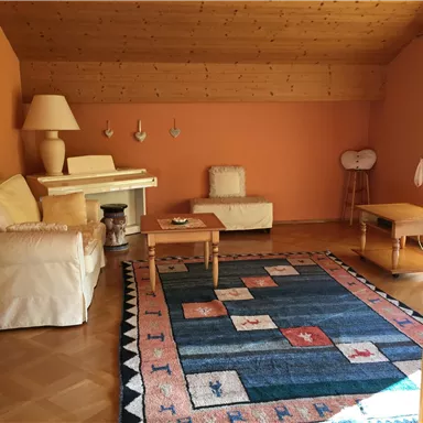 Ein gemütliches Wohnzimmer mit orangen Wänden und Holzbalkendecke. Es gibt ein bequemes Sofa, einen Couchtisch und einen bunten Teppich.