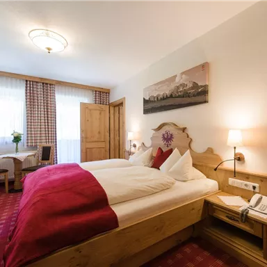 Ein gemütliches Schlafzimmer mit einem Doppelbett und roten Akzenten. Fenster mit viel Tageslicht und ein kleiner Tisch mit Stühlen im Hintergrund.