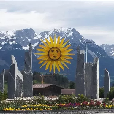 Eine große Sonne aus Holz steht in der Mitte einer Blumenrabatte, umgeben von steinernen Säulen. Im Hintergrund sind schneebedeckte Berge zu sehen.