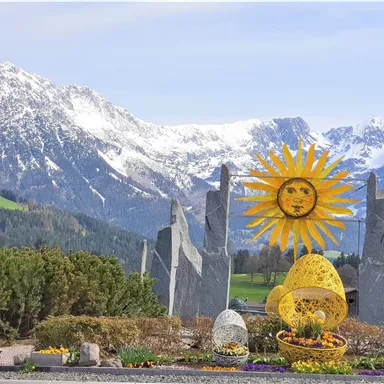 A colorful sun sculpture stands in front of snow-covered mountains. Surrounded by flowers and green bushes, the scene conveys a cheerful atmosphere.