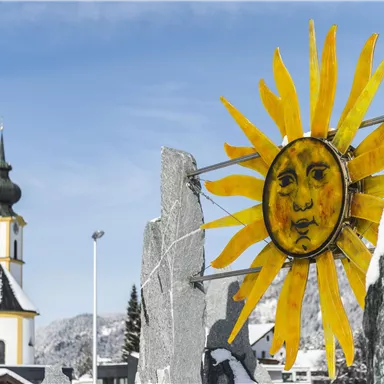 Ein sonniges Kunstwerk mit einem lachenden Gesicht steht vor einer verschneiten Landschaft. Im Hintergrund ist eine Kirche mit einem grünen Turm zu sehen.