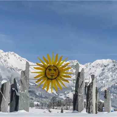 Ein winterlicher Bergblick mit einer sonnensymbolisierten Skulptur aus großen Steinen. Die schneebedeckten Berge im Hintergrund sorgen für eine beeindruckende Kulisse.