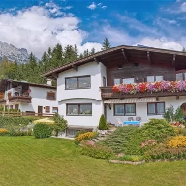 Ein modernes Haus umgeben von einem gepflegten Garten mit bunten Blumen. Im Hintergrund sind Berge und ein blauer Himmel zu sehen.