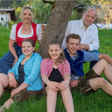 Eine Familie sitzt entspannt im Gras unter einem Baum. Sie tragen traditionelle bayerische Trachten und lächeln freundlich in die Kamera.