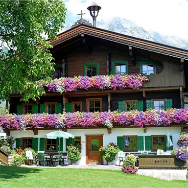Ein schönes alpenländisches Haus mit bunten Blumen auf dem Balkon. Im Hintergrund sind grüne Wiesen und Berge zu sehen.