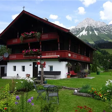 Ein traditionelles Alpenhaus mit roter Fassade und Blumenkästen. Im Hintergrund sind majestätische Berge und eine grüne Wiese zu sehen.