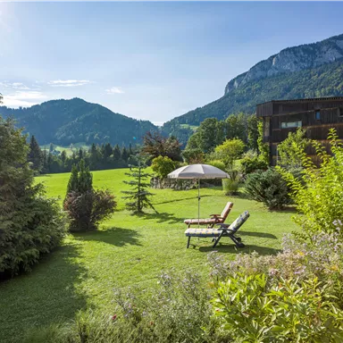 A beautiful garden with loungers and a sunshade. In the background, green mountains and a clear sky are visible.