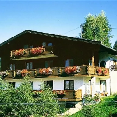 Ein gemütliches Holzhaus mit Balkonen und bunten Blumen. Es liegt in einer grünen Umgebung und hat einen klaren blauen Himmel im Hintergrund.