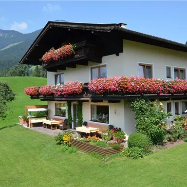 A charming house with blooming balcony flowers in a green environment. Gentle hills and mountains are visible in the background.