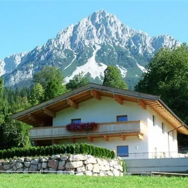 Ein modernes Haus mit einem Holz-Balkon, umgeben von Bäumen und einem gepflegten Garten. Im Hintergrund erhebt sich ein majestätischer Berg unter klarem Himmel.