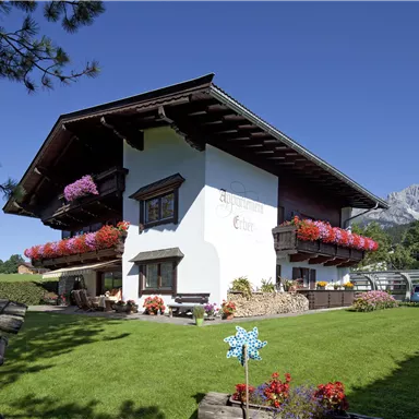 A charming white house with blooming balcony boxes and a well-kept green lawn. In the background, impressive mountains and a clear blue sky can be seen.