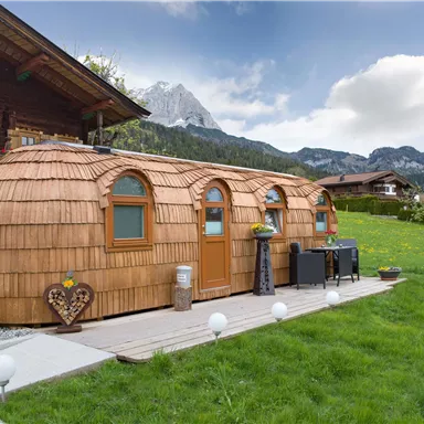 A modern wooden house with a unique, round design stands in a green environment. In the background, picturesque mountains and a clear sky can be seen.