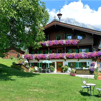 Ein charmantes Holzhaus mit bunten Blumenbalkonen und einem gepflegten Garten. Die Sonne scheint und umgeben von einer malerischen Berglandschaft.