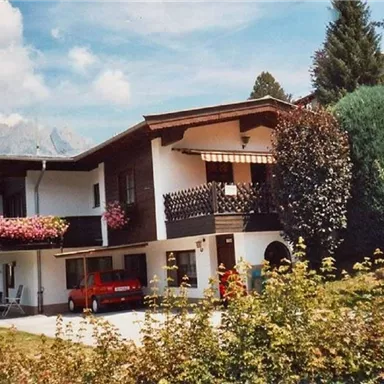 Ein charmantes zweigeschossiges Haus mit Balkonen und bunten Blumen. Umgeben von grünen Bäumen und Bergen im Hintergrund.