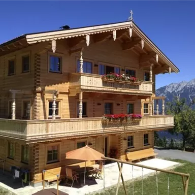 A modern wooden house with several balconies and flower boxes. In the background, impressive mountains and a clear blue sky can be seen.