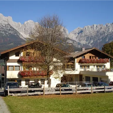 Ein hübsches Gebäude in den Bergen, umgeben von saftigem Grün und mit blühenden Balkonpflanzen. Im Hintergrund sind majestätische Felsen zu sehen.