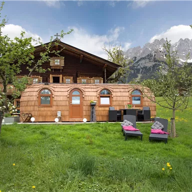 A cozy wooden cabin in Alpine style surrounded by green meadows and mountains. In front of the cabin, there are deck chairs and a beautiful garden.