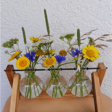 Eine Holzvase mit drei Glaskolben, gefüllt mit bunten Blumen. Die Blumen umfassen gelbe, blaue und grüne Blüten und verleihen dem Arrangement eine frische Note.